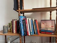 Books on Arrow and Avro aircraft, some aviation history, and Canadian history books arranged on a wooden bookshelf. Includes 'Aircraft of the World' and 'Avro Canuck & Avro Arrow' books plus framed airplane artwork.