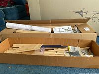 Open cardboard boxes containing rolled plans, packets of wood strips and small hardware bags