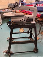 Front view of Dremel 16-inch scroll saw on black wheeled metal stand showing the body, worktable, and power cord.