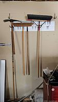 Four tools hung on a garage wall including broom with black bristles, wooden rake, wooden broom with light bristles, and an axe with wooden handle