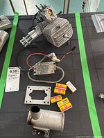 Photo showing the engine, muffler, six boxed spark plugs, ignition module, and mounting plate on a dark surface with green tape.