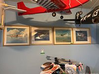 Four framed vintage aircraft prints hanging on wall under hanging model airplanes, each showing different vintage planes in flight.
