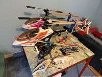 Three assembled RC helicopters on table, two with matching white/orange/pink canopies and one smaller unit without canopy fitted.