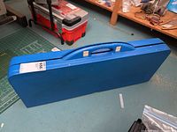 Closed blue plastic carrying case for folding picnic table with handle, shown from side profile.