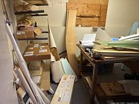 Overview of wooden boards, foam panels, boxed components, and rolled plans stored against wall