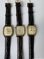Three vintage men's wristwatches with rectangular gold-tone cases and black/dark brown textured leather straps on white background.