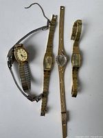 Four vintage women's wristwatches arranged on a white surface - one silver-tone with round face, three gold-tone with rectangular faces and various band styles