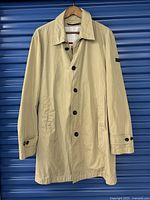 Beige men's trenchcoat on hanger, front view showing buttons, collar, pockets, and sleeve cuff straps.