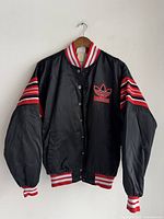 Front view of black vintage Adidas varsity jacket featuring red and white striped collar, waistband and cuffs, with red stripes on sleeves and embroidered Adidas logo on chest.