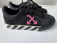 Side angle view of black Off White sneaker showing pink arrow logo and striped sole.