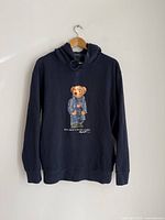 Front view of navy blue Polo Ralph Lauren hoodie featuring embroidered bear graphic with American flag and denim jacket on front. Hanging on wooden hanger against white wall.