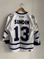 Back view of white hockey jersey showing 'SUNDIN' name and number 13 in blue with blue stripes on sleeves and bottom hem.