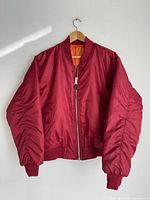 Front side view of the vintage Rothco MA-1 flight jacket hanging on a wooden hanger showing red nylon exterior, ribbed cuffs, waistband, and full zipper closure.