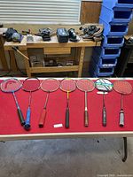 Seven badminton rackets laid out on a red table with various colors and grips visible.