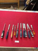 Photo showing set of 11 woodworking chisels with a mix of black and white rubberized handles, blue plastic handles, wooden handles, and translucent yellow handles arranged on a red surface.