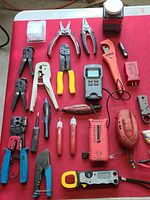 Overview of electrician tools including wire strippers, crimpers, circuit tracker, clamp meter, testers, and adapters laid out on red surface