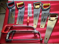 Seven different hand saws, including backsaws and hacksaws, laid flat on red surface.