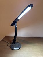 Black LED desk lamp turned on, placed on wooden table showing bright light emission.