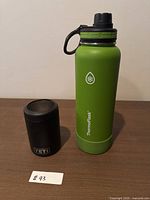Green Thermoflask water bottle and black Yeti can holder side by side on wood surface.