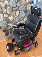 Side angled view showing black padded seat, armrest with joystick control, footrest, and red rear fender.