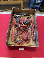 Box with a large assortment of screwdrivers, plastic handles mostly red and black, various sizes.
