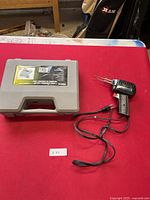 Full view of the soldering iron and the grey carrying case containing the soldering kit set on a red surface