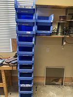 Vertical stack of 11 blue plastic bins arranged on the floor next to a workbench and wall with a window, showing their general size and condition.