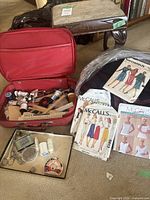 Photo of open red sewing kit case filled with assorted sewing items, metal tray with thread spools and scissors, vintage sewing patterns laid out nearby, wooden footstool behind.