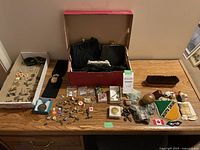 Full overview of collection showing vintage bags in box, boxed watches, assorted pins, badges, keychains, cufflinks, and miscellaneous small items on a wooden table.