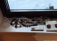 Multiple necklaces laid out on a surface including beaded black, cream, and mixed stone-like beads. Red plaid stud earrings on GITANO brand card. Metallic thermometer-style pendant in silver tone.