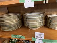 Stacks of white porcelain dinner plates and smaller plates with gold trim and monogram on shelf. Some minor chips noted.