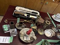 Wide view of antique mixed metal and brass candle holders, ceramic candle holder, small decorative plates, antique flatware, and candle accessories on table.