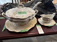 Overview of porcelain soup serving bowl with lid, matching underplate, and ladle beside porcelain gravy boat set with its underplate and ladle on a table surface.