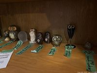 Nine owl figurines displayed on wooden shelf showing diverse materials and styles.