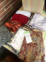 Six folded scarves in various colors and patterns, including red, brown, black, white, gold, and purple, placed against a brick and tiled backdrop.