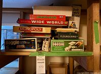 Multiple vintage board games stacked on a shelf, including Monopoly, Wide World, Canada-opoly, Boggle, and Floundering. Visible wear and discoloration noted on game boxes.