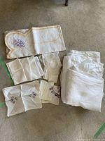 Set of white linens including several folded tablecloths, embroidered napkins with purple floral motifs, and crochet doilies on carpet floor.