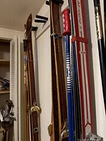 Photo showing a set of vintage wooden skis and several ski poles with different handles stored upright against a wall.