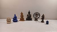 Long shot showing all seven assorted Buddha, goddess, and Ganesh statues lined up on a flat surface against a plain background.