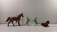 Four decorative horse figurines arranged in a row: brown hollow metal horse with ornate saddle and chain reins, two green plastic horses standing on rear legs, and a brown resin horse lying down.