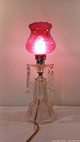 Table lamp lit showing cranberry colored hobnail glass shade emitting pink light, clear cut glass base with dagger-shaped crystal pendants.