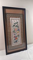 Overall view of framed embroidered tapestry, showing dark wooden frame and beige matting