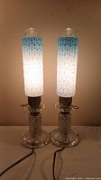 Both lamps illuminated, showing embossed blue glass bullet shades and clear pressed glass bases