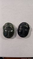 Top view of two hand-carved stone scarab beetles showing detailed carved body segments with green and black color variations, rough edge visible on the green scarab.