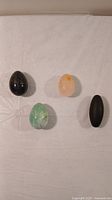Four polished stone items including two crystal eggs (black, green), one peach crystal egg chipped at bottom, and one black oblong stone arranged on a white surface.