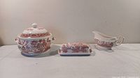 Front view of Churchill porcelain serving pot with lid, butter dish with lid, and gravy boat arranged in a row on white surface, showing red transferware decoration on cream porcelain.