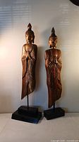Front view of two standing Buddha statues with dark finish on black bases. Taller statue has wall hanging attachment and some paint chipping.