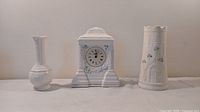 Three white Belleek porcelain pieces on table: Claddagh bud vase, Cashel clock, Castle vase