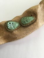 Pair of antique gilt silver shoe clips with hand-carved turquoise stones where one turquoise shows noticeable damage and the silver frames have patina and tarnish, resting on a wooden display.