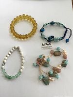 Four beaded bracelets laid flat on white background: yellow round bead stretch bracelet, blue and green knotted bracelet with adjustable closure, white marbled bead stretch bracelet with green beads, multi-stone bracelet with turquoise, rose quartz-like stones and pearls with knotted string design.